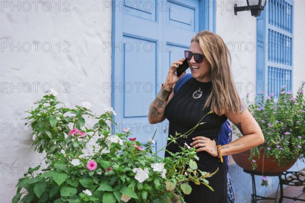 A joyful middle-aged woman with straight hair and a curvy Latin figure chats on the phone in front of a vibrant blue colonial door. She exudes charm and confidence in a stylish black dress and sunglasses.