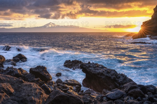 Sunrise on the beach at San Marcos. Rocks, sea and the sun. Tenerife and the Teide in the background. Bright clouds in the sky. La Gomera, Canary Islands, Spain, Europe