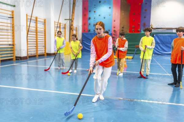 Group of elementary school students wearing orange and yellow vests playing floorball in a gym class, learning teamwork and sportsmanship