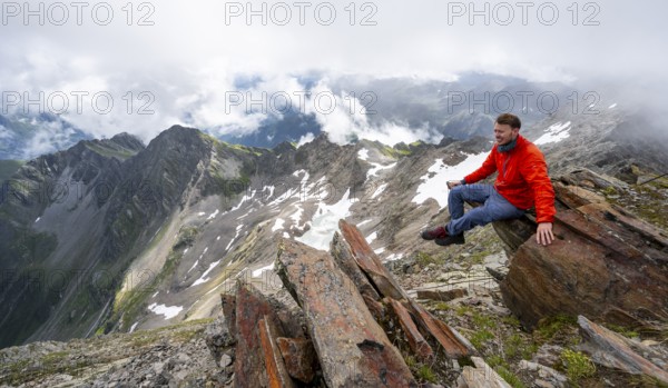 Mountaineer sitting on a rock, cloudy mountain landscape at the summit of Lasörling, view to Berger Kees and mountain peak Schober, Lasörling Group, Hohe Tauern National Park, East Tyrol, Tyrol, Austria, Europe