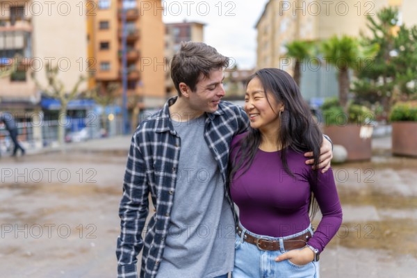 Joyful multiethnic couple shares a happy moment outdoors, embracing and smiling at each other, surrounded by urban buildings and greenery