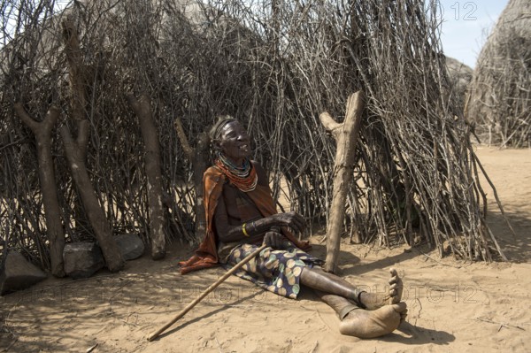 Old woman sitting in front of a fence made of wooden sticks and small tree trunks, Karo ethnic group, Southern Omo Valley, Ethiopia, Africa