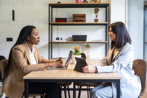 Two businesswomen are working together on a project using a laptop and tablet in a hotel lobby during a business trip