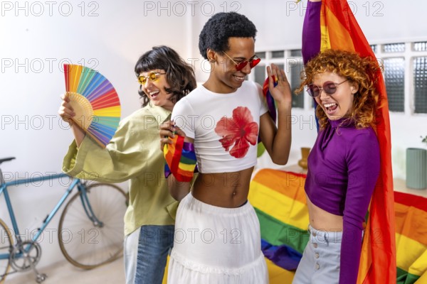 Happy multi-ethnic and gender diverse friends celebrating pride party at home wearing sunglasses and using hand fan and rainbow flags while dancing