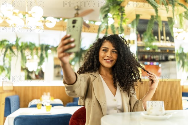 Sensual and happy latin woman taking selfie while touching her long curly hair sitting alone in a cafeteria