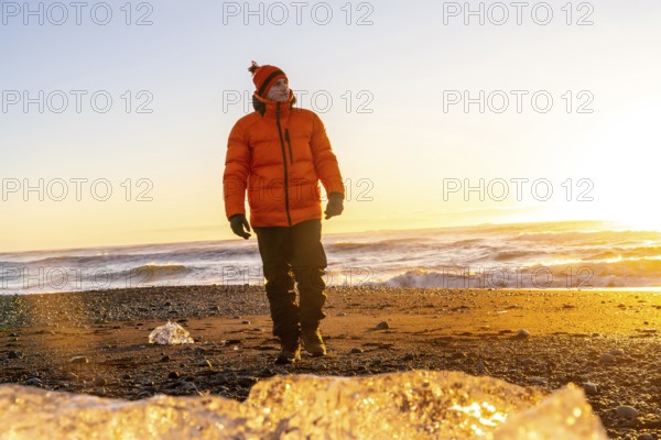 A tourist in an orange jacket walking at sunrise on Diamond Beach Iceland in winter