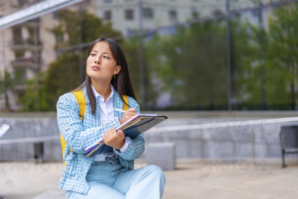 Portrait three quarter length of a beauty Asian student writing notes sitting on the campus looking up