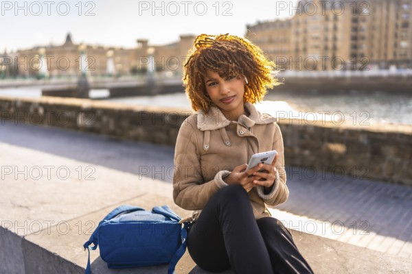 Beauty latin female student using smartphone sitting next to rucksack on the city during sunset
