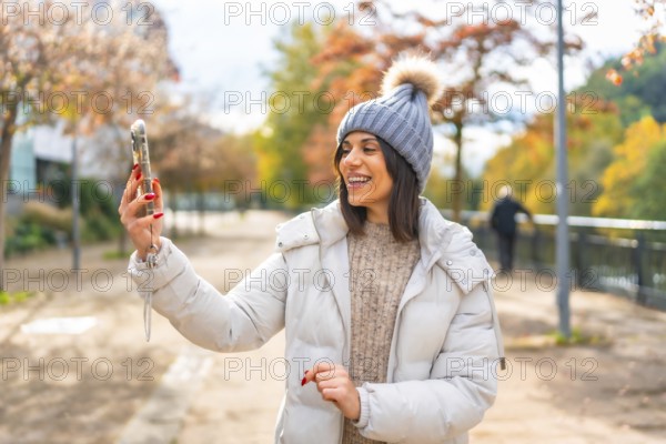 Woman wearing warm clothes and woolly hat smiling while talking during video call with mobile phone walking along a city park