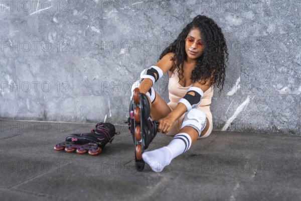 Latin beauty woman putting on inline skates, sitting in front of an urban wall, wearing pink sportswear