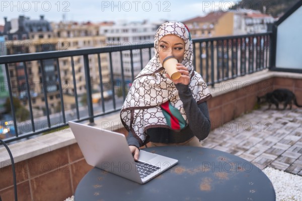 Arab girl with a white veil at the computer on the terrace of a coffe shop, doing work. Modern Arab young man with new technologies