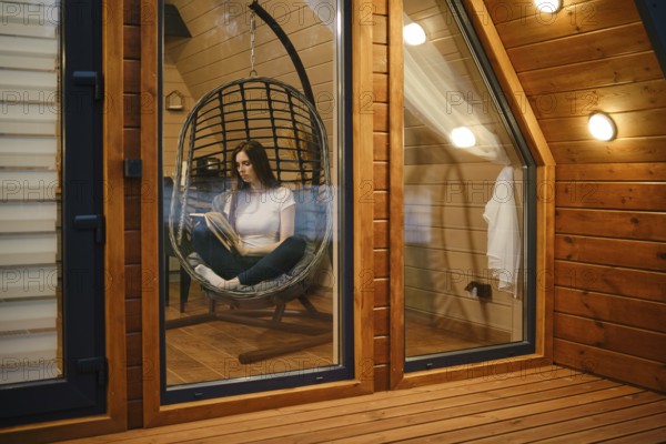 Cute woman sitting in a hanging swing chair and reading a book in the evening