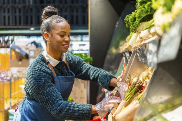 Smiling saleswoman wearing gloves arranging fresh organic asparagus on display in a supermarket