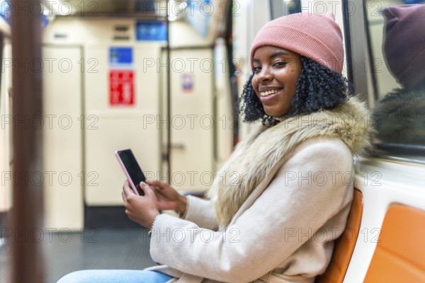 Young black woman wearing winter clothes and using mobile phone while sitting and traveling by subway