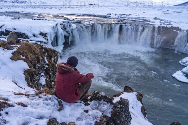 A man hiker sitting on the frozen Godafoss waterfall at sunset in winter, Iceland, Europe