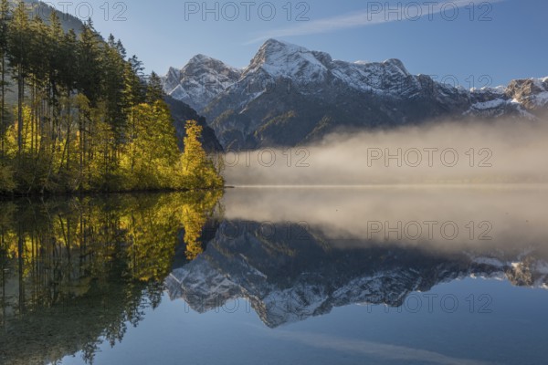 Lake Almsee in autumn with a forest and mountains reflected in the lake. A slowly dissipating fog moves over the lake. Blue sky