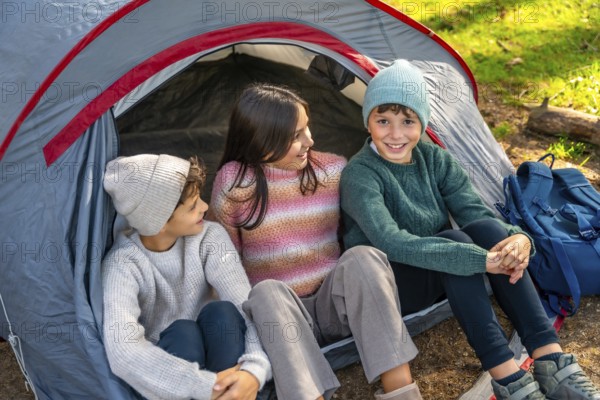 Three caucasian kids enjoying camping day in the forest sitting inside the tent and smiling