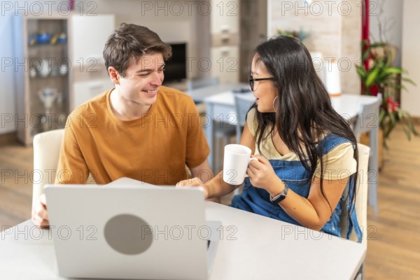 Young multiethnic couple smiling and looking at each other while using laptop and drinking coffee, sitting at table in their modern apartment