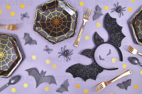 Halloween party flat lay with spider web plates, costume bat headband, spiders and confetti on violet background