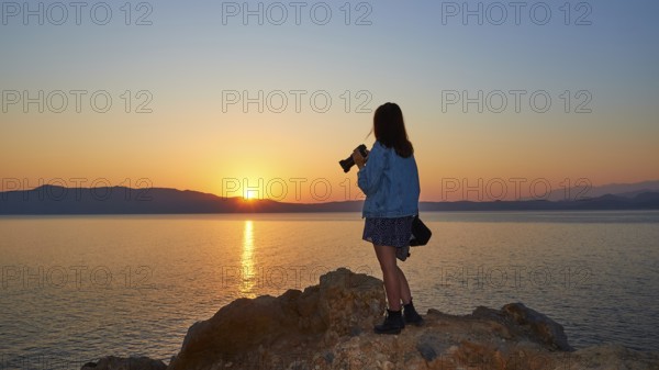 Sunrise, sun reflection on the sea, orange-blue cloudless sky, girl from behind with jeans jacket and camera in hand, Rodopou peninsula, Gramvoussa peninsula, Pirate Bay, Balos, Tigani, West Crete, island of Crete, Greece, Europe
