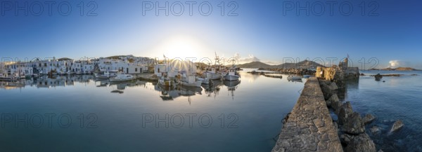 Panorama, fishing boats in the harbour of Naoussa at sunset, reflected in the sea, sun star, ruins of the Venetian castle on the right, white Cycladic houses, Naoussa, Paros, Cyclades, Greece, Europe