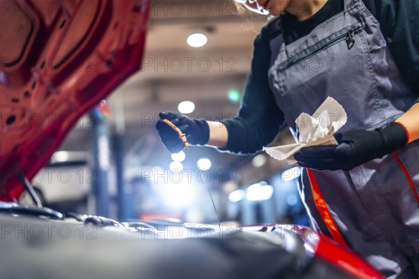 Professional female mechanic inspecting engine oil level with dipstick and rag in a car repair shop
