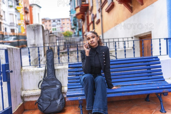 African american woman sitting on a city blue bench talking to the mobile with a guitar next to her