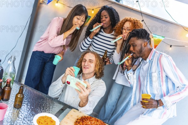Happy caucasian man taking selfie while friends blowing streamers during birthday rooftop party