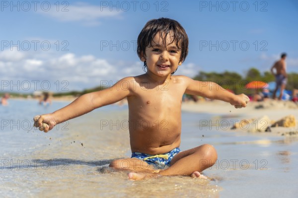 A young boy is sitting in the sand on a beach, smiling and looking out at the water