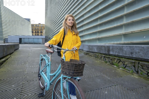 Young woman wearing a yellow raincoat and backpack walks with her bicycle in a modern city setting