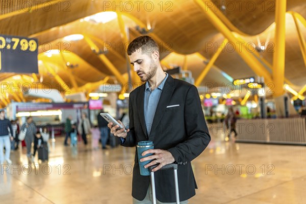 Businessman waiting for his flight, using smartphone and holding reusable coffee cup at the airport