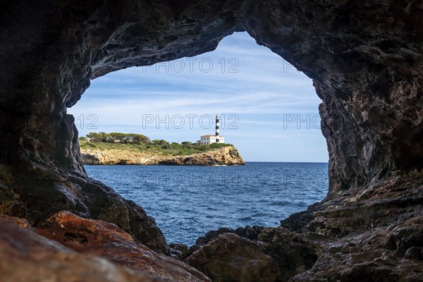 Portocolom lighthouse framed by the rocky opening of the arc natural cave, with blue sea and sky in the background, in mallorca, spain