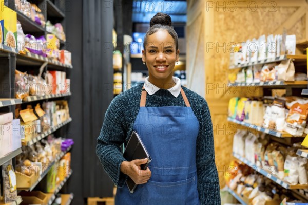 Confident saleswoman smiling and holding tablet, working in organic supermarket with shelves full of natural and healthy products