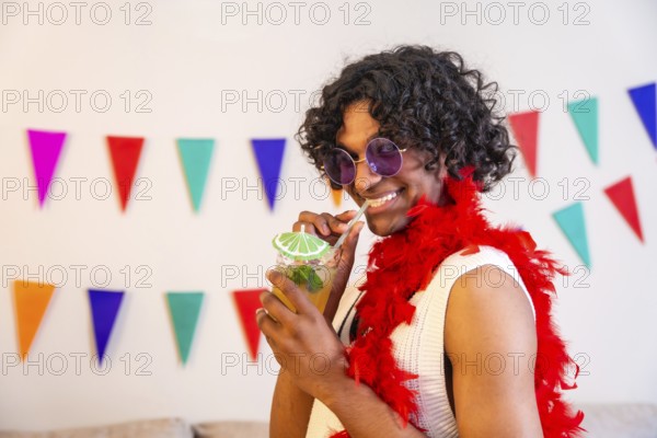 Portrait of a happy brunette gay man drinking cocktail next to colorful garlands at home