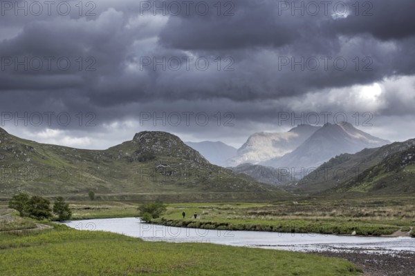Two walkers walking on path along river and rain clouds forming in Wester Ross, Scottish Highlands, Scotland, UK
