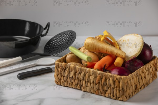 A woven basket full of vegetables such as soup vegetables, potatoes and onions next to kitchen utensils
