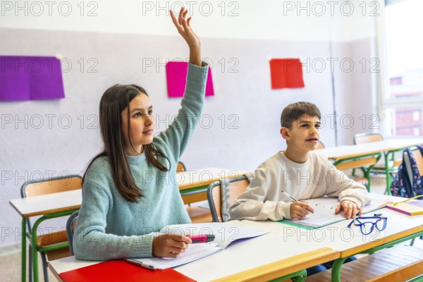Girl raising hand to answer a question during class in elementary school while her classmate is taking notes