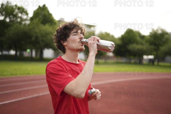 Thirsty caucasian young amle athlete drinking water in a reusable bottle after training in a running track