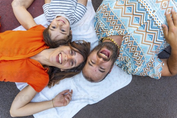 A family of three, a man, a mother and a son, are laying on a blanket on the ground. The man is wearing a blue and white shirt with a pattern of diamonds. The children are smiling