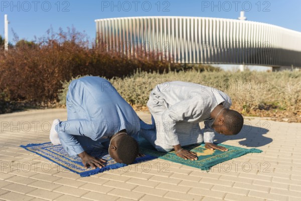 Two senegalese men wearing traditional dashiki clothing are kneeling on prayer rugs, performing salat during the holy month of ramadan