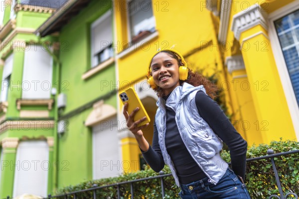 Low angle view photo of a cool afro young woman in urban clothes listening to music next to colorful houses