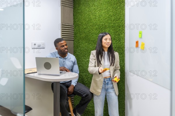 Two managers are discussing a project using sticky notes on a whiteboard in a modern coworking office with a green wall