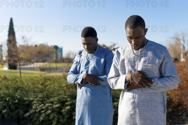 Two young senegalese muslim men wearing traditional dashiki clothing are performing salat prayer outdoors in a park during the holy month of ramadan