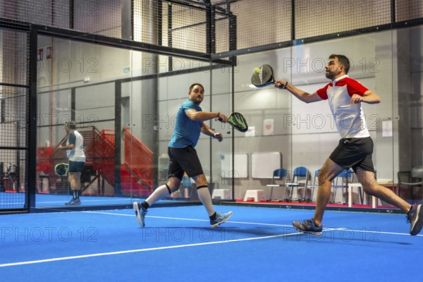 Mature caucasian male friends playing paddle tennis energetically in a blue indoor court