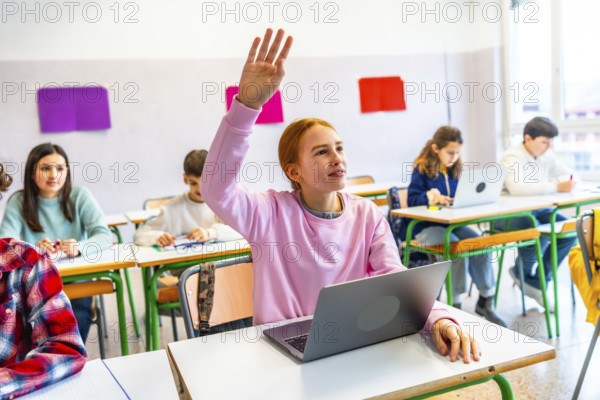 Smart student raising hand to answer teacher's question in modern classroom with laptops and focused classmates