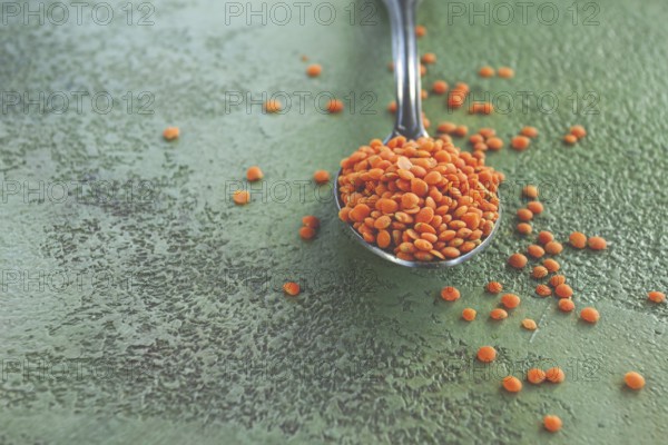 A spoon holds vibrant red lentils, with more lentils scattered around on a textured green surface. The image highlights the color and texture of the lentils