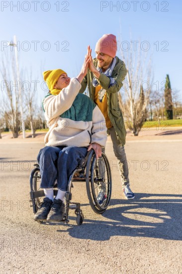 Two friends celebrating something with a high five in a park, one of them using a wheelchair, concept of disability and friendship