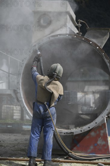 A man weating full safety gear uses ground glass to sandblast the steel casing of a meat drying machine for a rendering plant