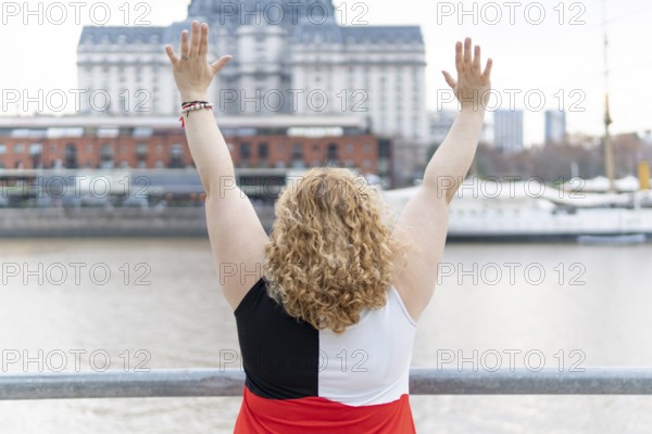 Plus size blonde woman on her back standing on the waterfront with her arms outstretched celebrating
