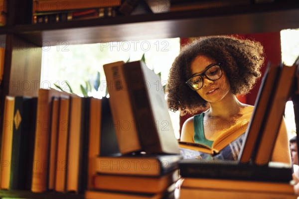 Happy African American female student studing in university library and looking for the book, wearing glasses